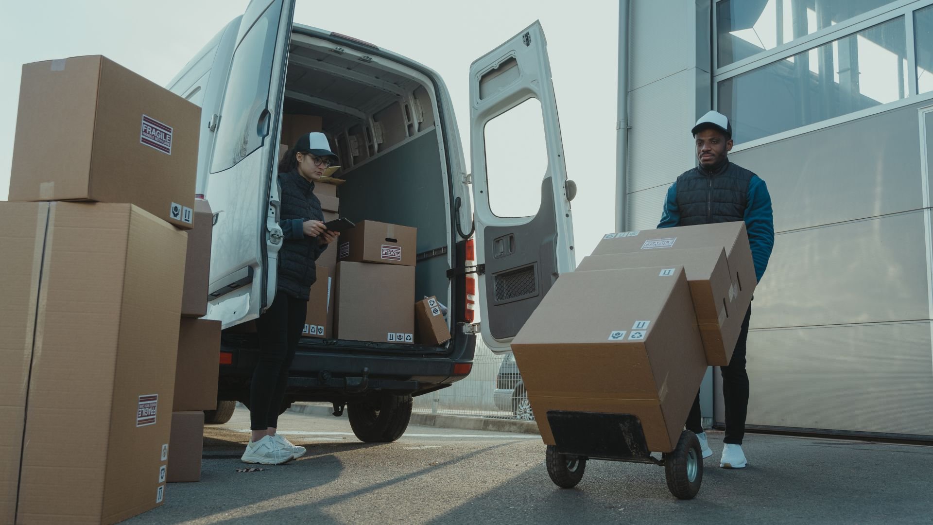 Delivery workers loading cardboard boxes into a white van using hand truck
